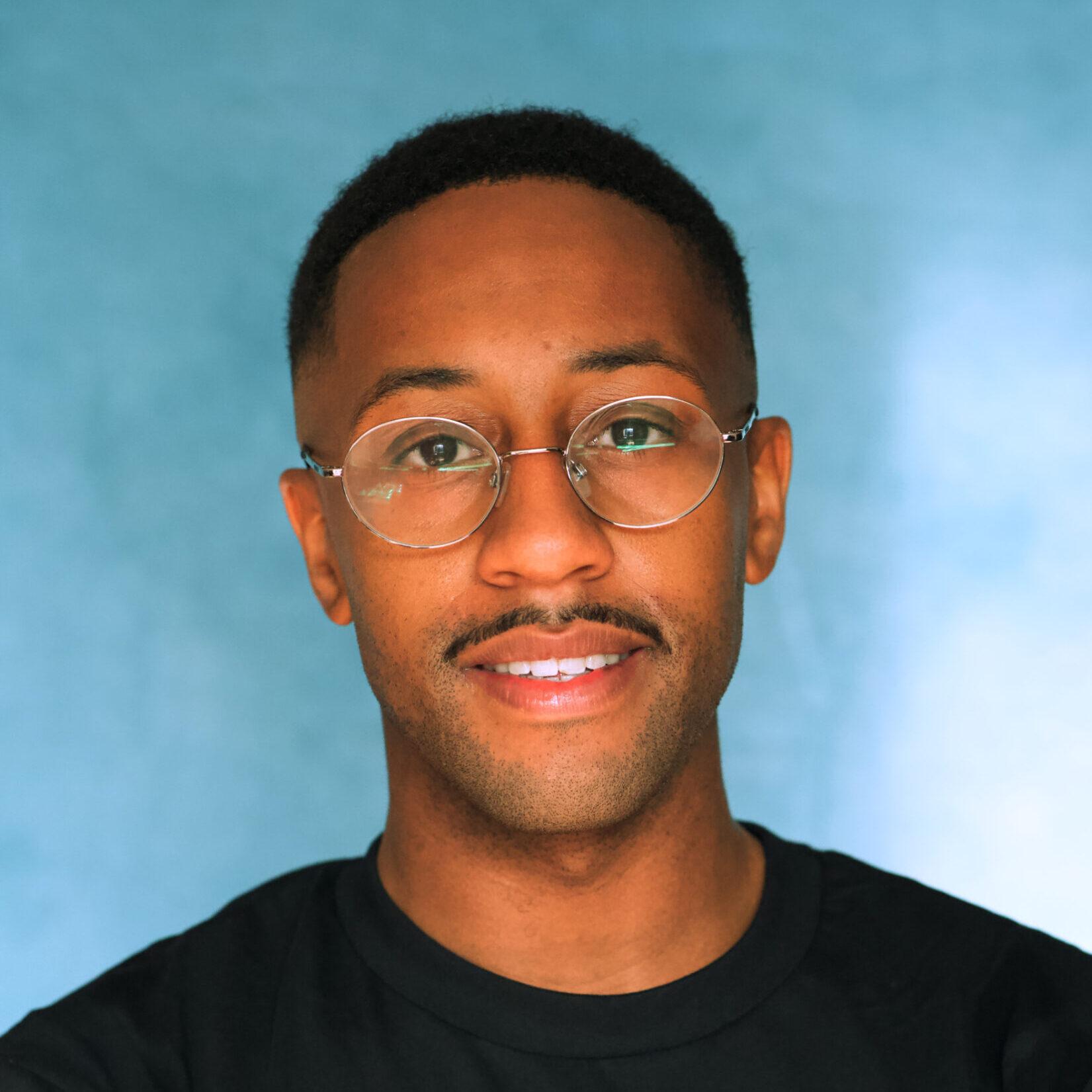 Tashan is a Speaker, coach and workshop facilitator at The Hobbs Consultancy who specializes in empowering underserved leaders to bring their authentic selves to the forefront. He is looking at the camera and wears a black top and metal framed glasses.